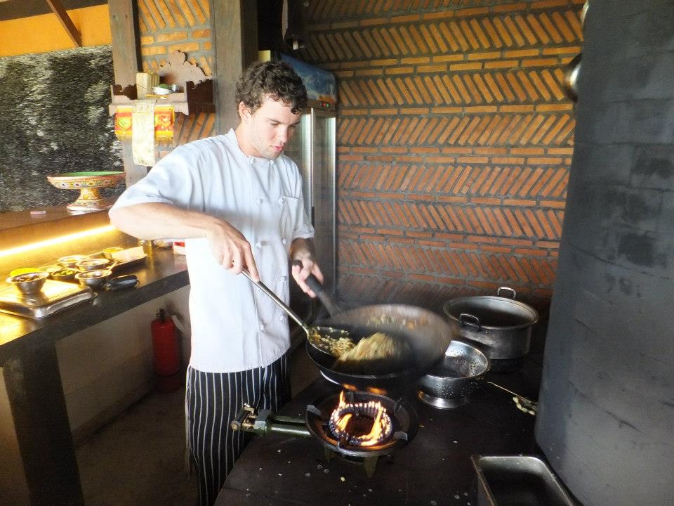 A chef in a white shirt and striped apron stir-fries ingredients in a wok over an open flame in a traditional kitchen setting.