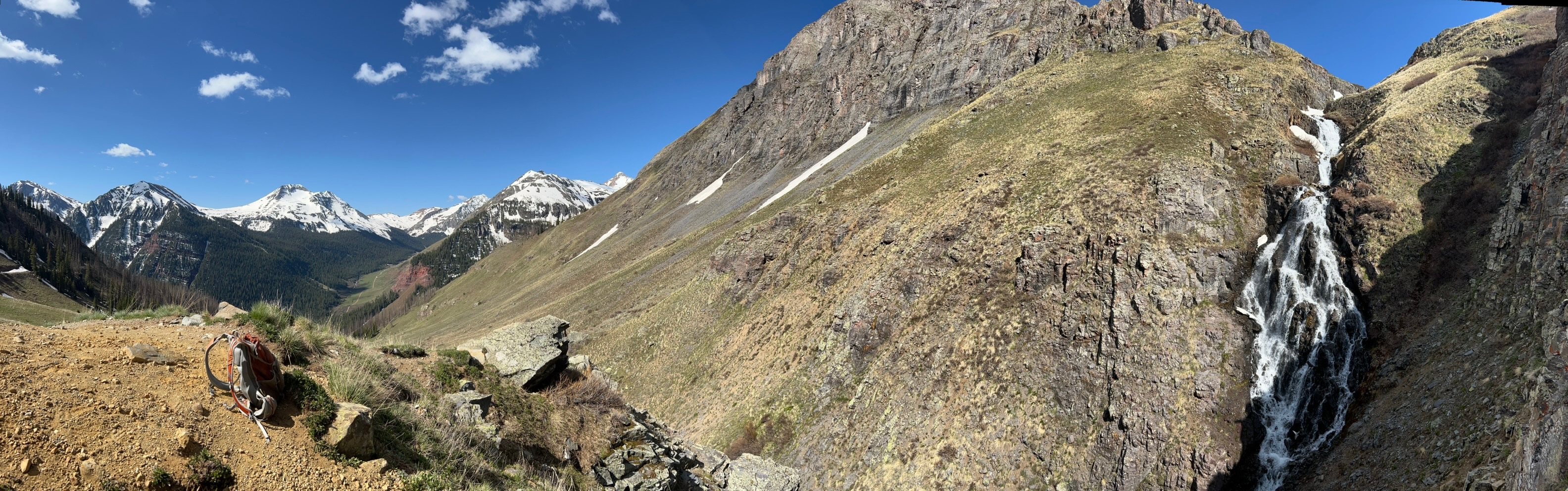 A mountainous alpine landscape with snow-capped peaks, a cascading waterfall, and a hiking backpack resting on rocky terrain under a clear blue sky.