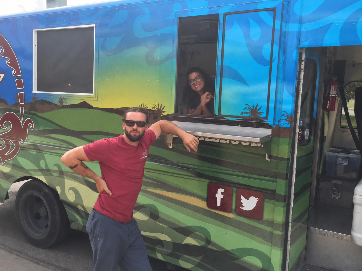 A man in a maroon shirt and sunglasses poses in front of a colorfully painted food truck with blue and green desert-themed artwork and social media icons.