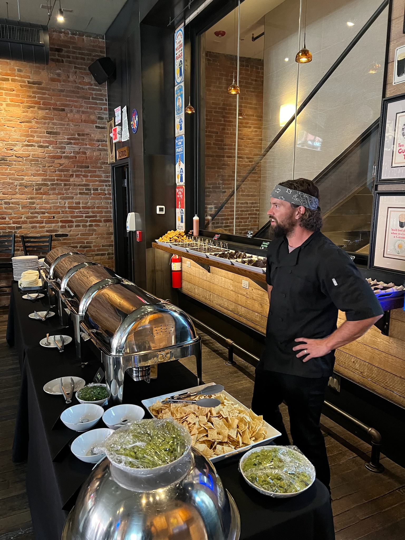 A man in black chef's attire stands beside a buffet table displaying multiple warming trays with various prepared foods and garnishes in a modern restaurant with exposed brick walls.