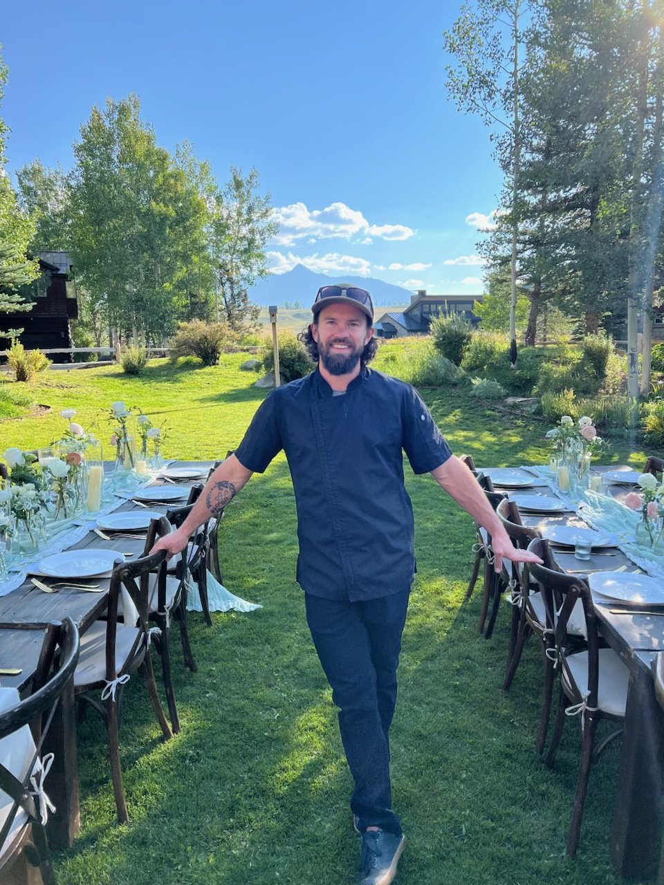 A smiling man in a dark blue chef's shirt and cap stands between two long outdoor dining tables set with place settings in a scenic mountain valley landscape.